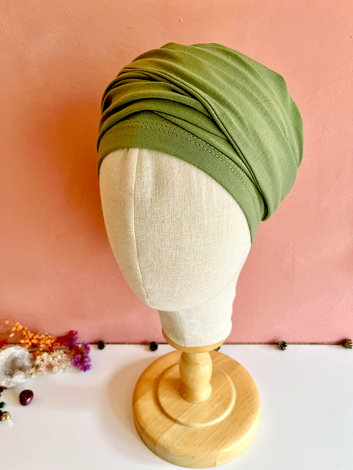 Green twist headwrap on beige mannequin head against a pink wall, displayed on a wooden stand.