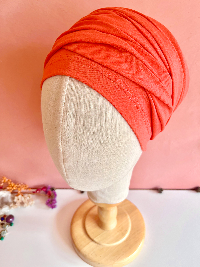 Orange turban wrap on a beige mannequin head against a pink wall, mounted on a wooden stand. Jewelry lies nearby in the corner.