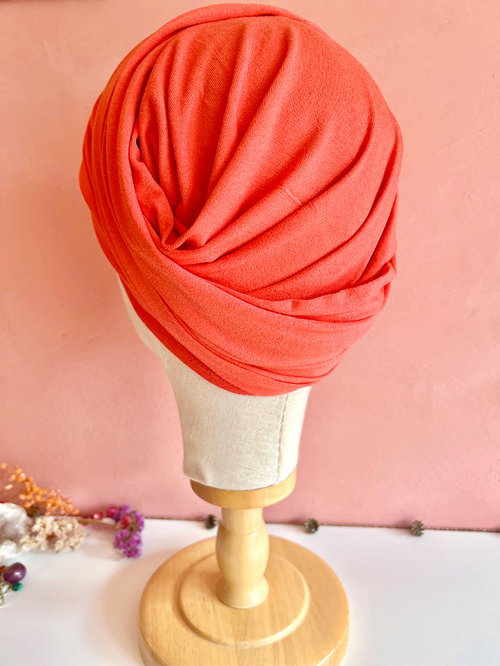 Coral red turban-style head wrap displayed on a mannequin head with a wooden stand against a pink wall.
