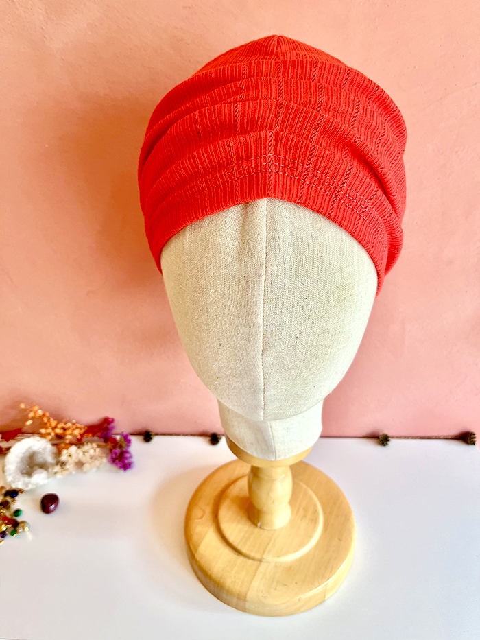 Red textured turban-style hat displayed on a beige mannequin head against a pink wall.
