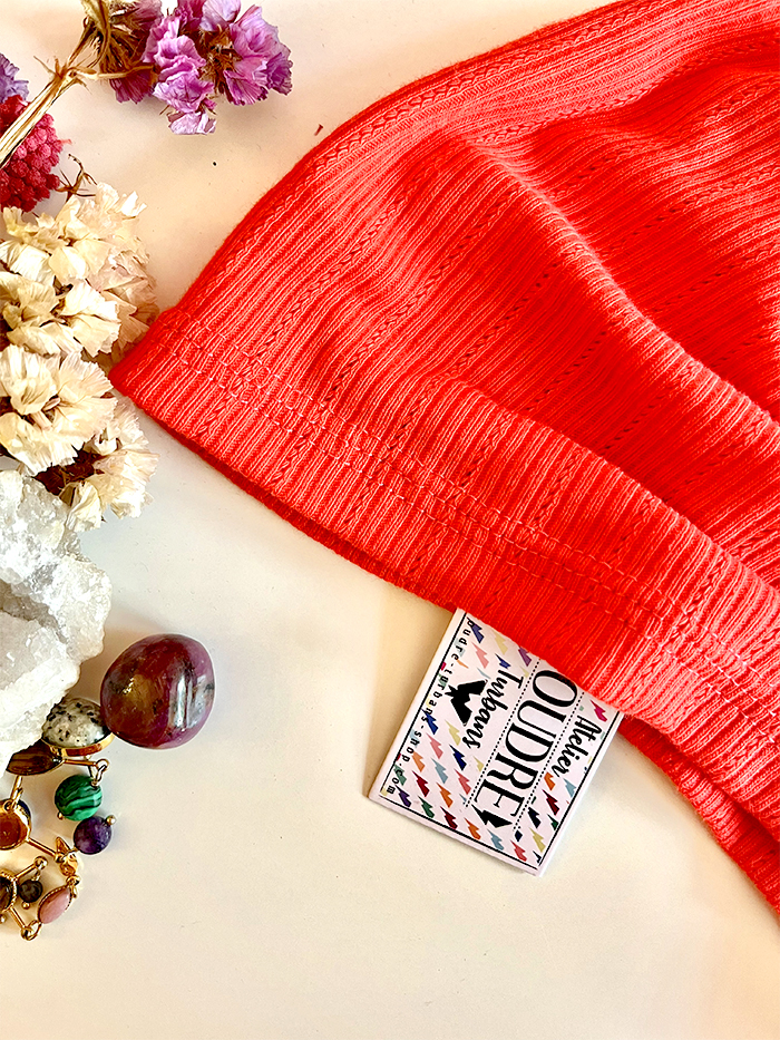 Flat lay of a bright red knit beanie surrounded by dried flowers, colorful jewelry, and a small patterned card on a light background.