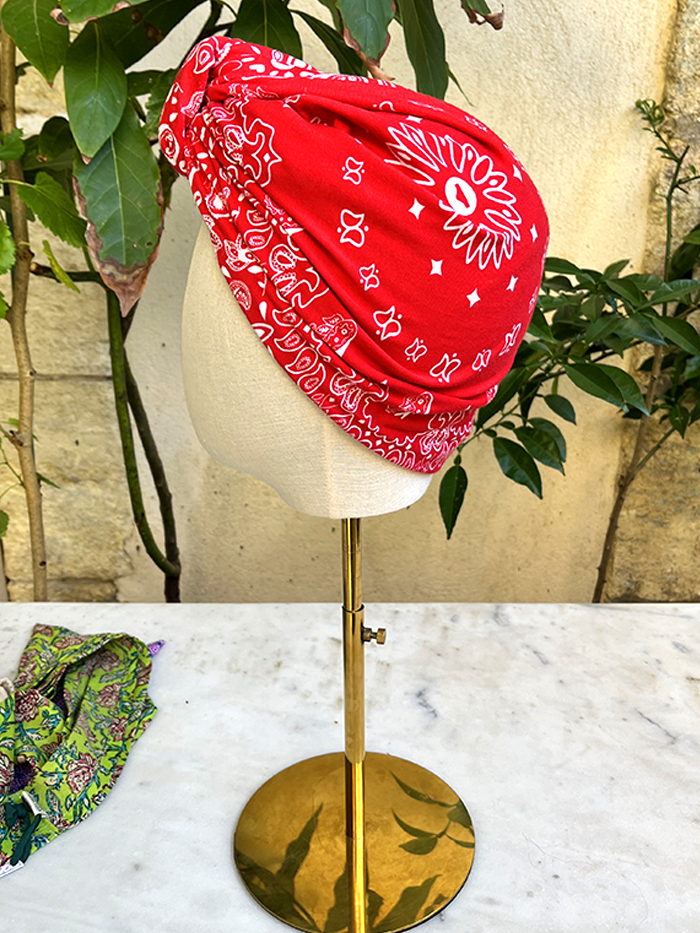 Red bandana with white paisley pattern wrapped around a beige head form on a gold-finish stand outdoors.