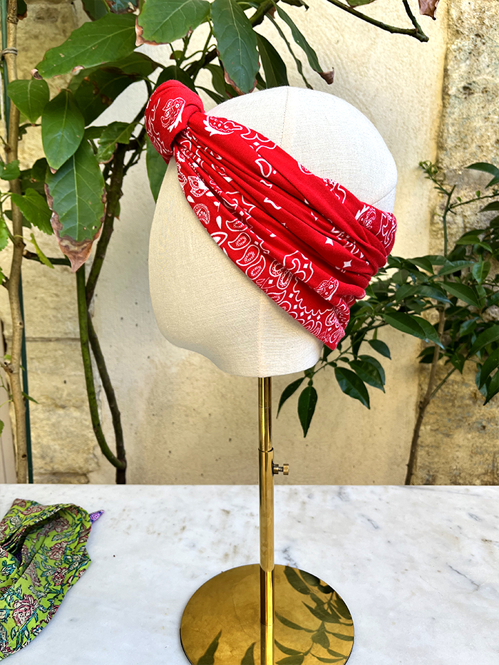Red bandana headwrap wrapped around a white mannequin head on a gold stand, outdoors with plants in the background.