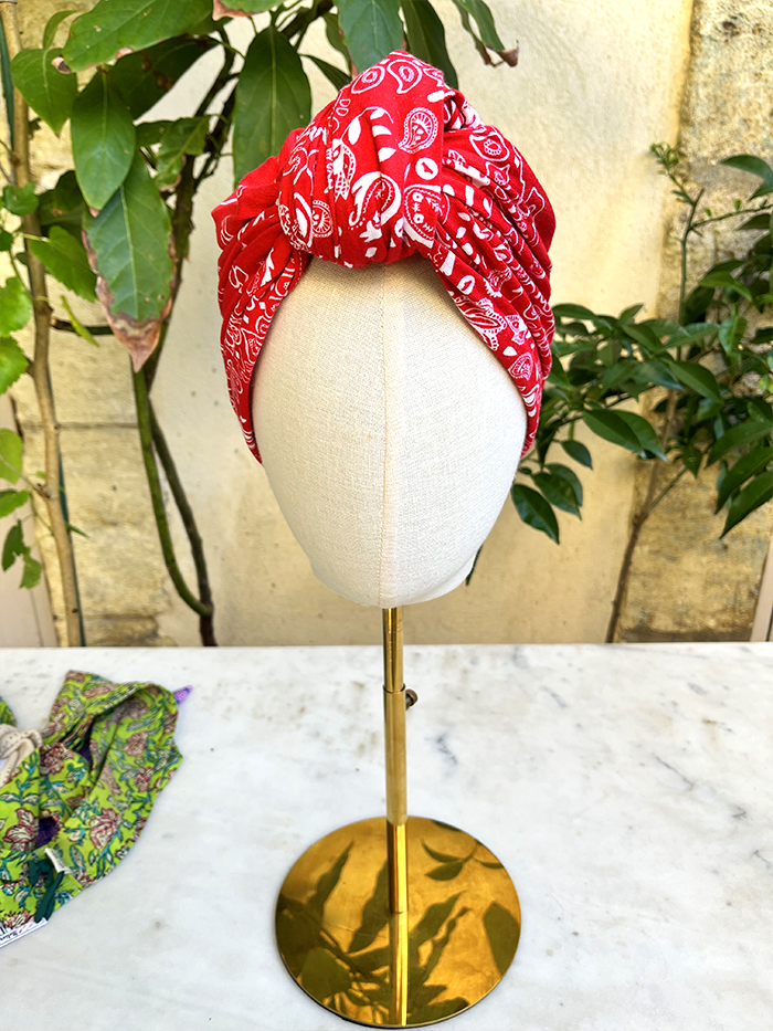 Beige mannequin head on a gold stand wearing a red patterned fabric head wrap outdoors.