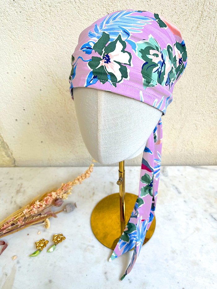 Pink tropical floral headscarf wrapped around a mannequin head on a brass stand, with a long tie hanging down the side.