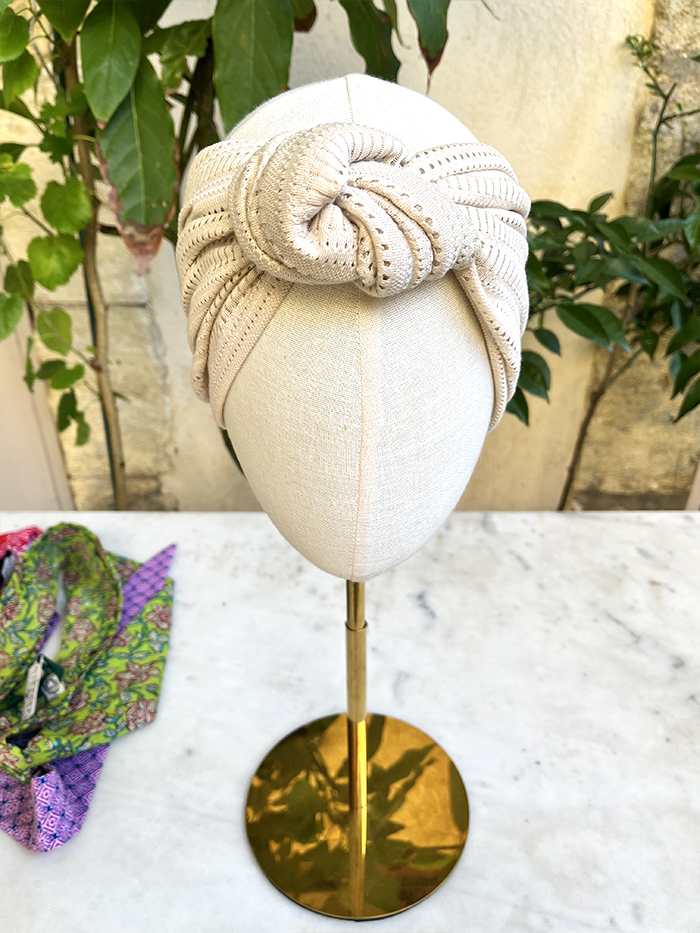 Beige knotted turban headwrap displayed on a dress form outdoors.