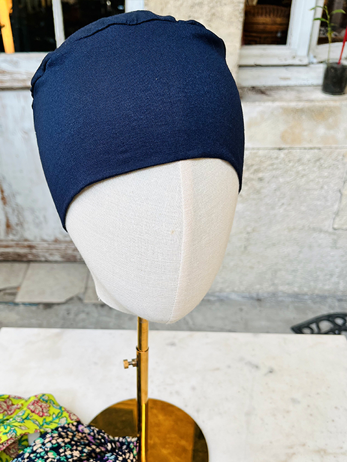 Navy blue fabric cap displayed on a dress-form mannequin head on a gold stand outdoors, with a stone wall in the background.