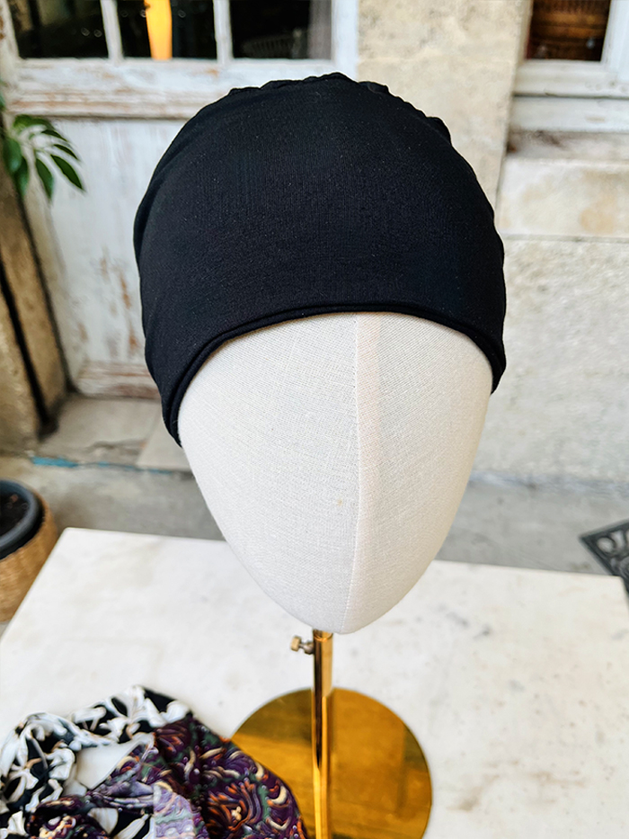 Black turban-style head wrap displayed on a beige mannequin head outdoors on a brass stand, with patterned scarf beneath it on a table.