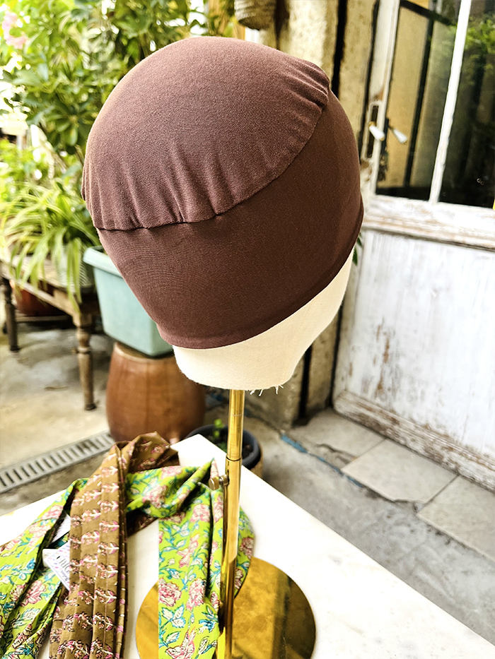 Brown fabric cap displayed on a beige mannequin head on a stand, with colorful scarves nearby in a shop setting.