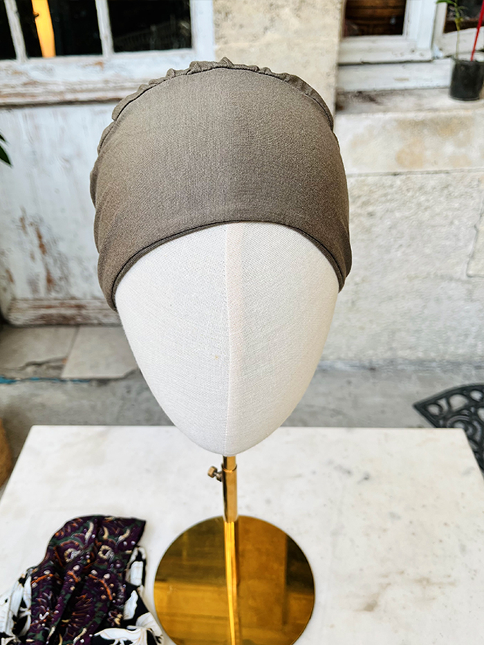 Beige head mannequin wearing a taupe fabric turban with a ruffled edge, displayed on a stand outdoors; a patterned scarf rests on the table nearby.
