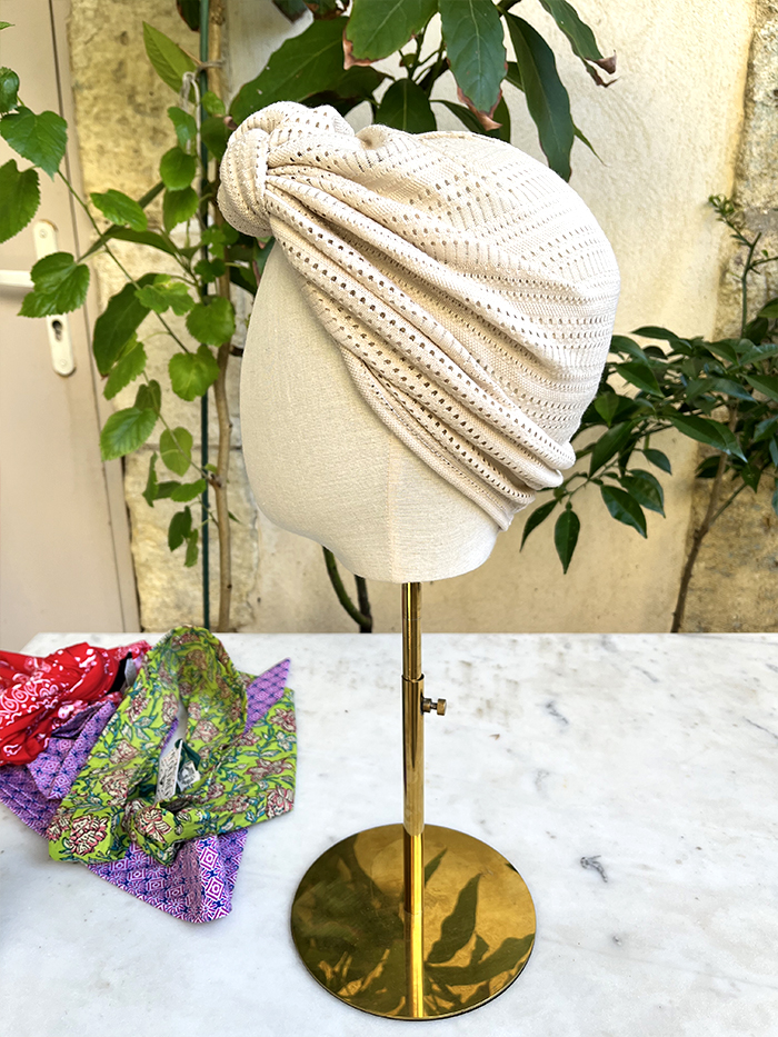 Beige knit turban shown on a dress-form head on a brass stand outdoors, with colorful fabrics nearby.
