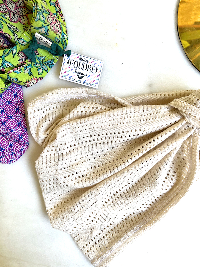 Beige knitted scarf with an eyelet pattern laid flat on a white surface, surrounded by a green floral scarf and other fabrics, with a small label nearby.