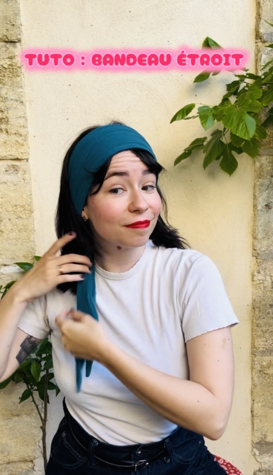 Woman wearing a teal headscarf/turban, white shirt, and red lipstick, adjusting a blue scarf outdoors against a stone wall with greenery nearby.