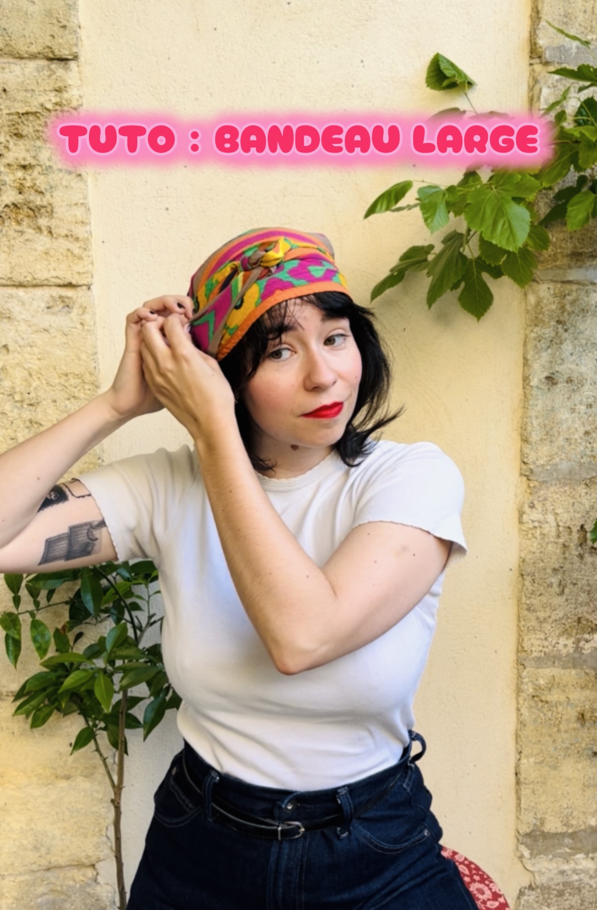 Woman fixing a colorful headscarf on her head, standing against a beige stone wall; pink text overlay reads 'TUTO : BANDEAU LARGE'